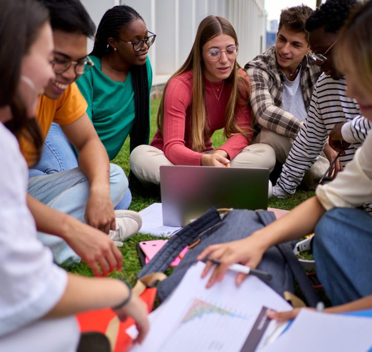 Vertical. Group of young international students sitting on the grass outside the university building. Friends classmates gathered happy studying together with workbooks and using laptop on campus
